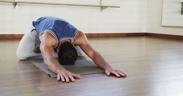 Man in athletic attire practicing child's pose on yoga mat inside fitness studio. Ideal for depicting mindfulness, relaxation, physical fitness, self-care, wellness sessions, and indoor activities.