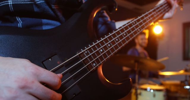 Focusing on hands as musician plays a black electric bass guitar with drummer in background in studio. Ideal for content related to music production, band rehearsals, musical education, and rock performances.