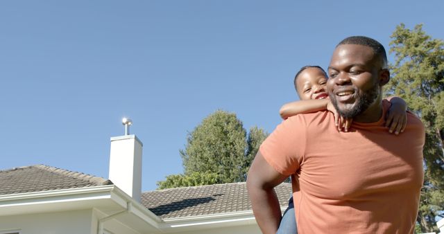 African American father giving piggyback ride to smiling daughter in the backyard of a home. Clear blue sky above and trees visible in background. Great for use in family-oriented advertisements, parenting blogs, bonding activities campaigns, and outdoor lifestyle promotions.