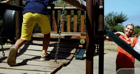 Child Climbing Wooden Bridge on Playground While Mother Watching and Smiling on Sunny Day