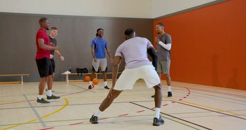 Multiracial men practicing basketball drills on indoor gym court with coach and clipboard