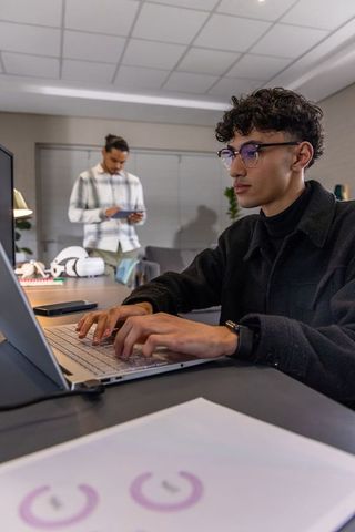 Young professional typing on laptop in modern office with coworker using tablet and vr gear