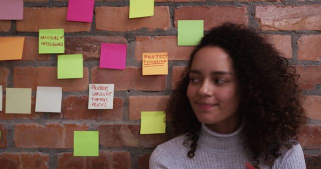 Young professional smiling while sitting next to a brick wall decorated with colorful sticky notes. Ideal for business, creativity, teamwork, and planning concepts. Perfect for illustrating office environments, brainstorming sessions, and collaborative workspaces.