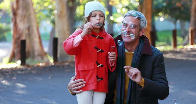 Grandfather and granddaughter spending quality time together in park blowing bubbles. Ideal for use in family bonding, grandparents' day, outdoor activities, playful moments, and autumn themed content.