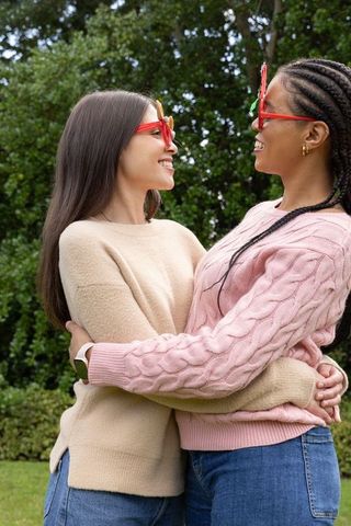 Multiracial women hugging and laughing outdoors wearing novelty glasses and sweaters