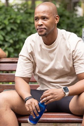 African american man sitting on park bench holding blue water bottle wearing smartwatch