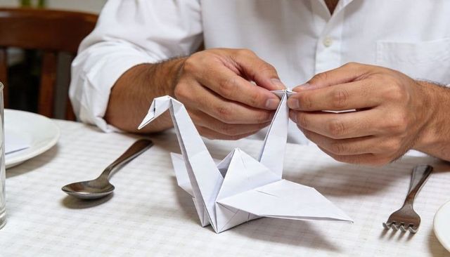 Man folding origami cranes at dining table, hands crafting delicate paper swans