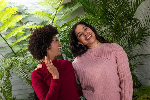 Two women laughing together in lush indoor garden wearing knit sweaters