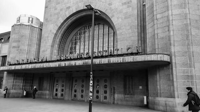 The image displays a classic railway station building photographed in black and white. The entrance features grand arched windows and vintage design elements. There are people in front of the building, adding a sense of scale and activity. This image is ideal for projects related to historical architecture, urban development, travel, and vintage themes.
