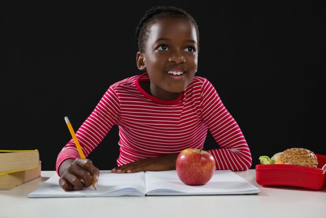 Young schoolgirl smiling while doing homework at desk with apple and lunchbox. Ideal for educational content, school promotions, and learning resources.
