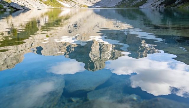 Alpine peak mirroring in crystal lake with cloud reflections and submerged pebbles