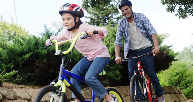 Father and daughter enjoying quality time riding bicycles together in a natural park. Both wearing helmets, they are smiling and experiencing the joy of a healthy outdoor activity. Ideal for use in advertisements promoting family time, healthy lifestyle choices, outdoor adventures, and cycling safety awareness. Suitable for use in parenting blogs, fitness magazines, and family-oriented product campaigns.