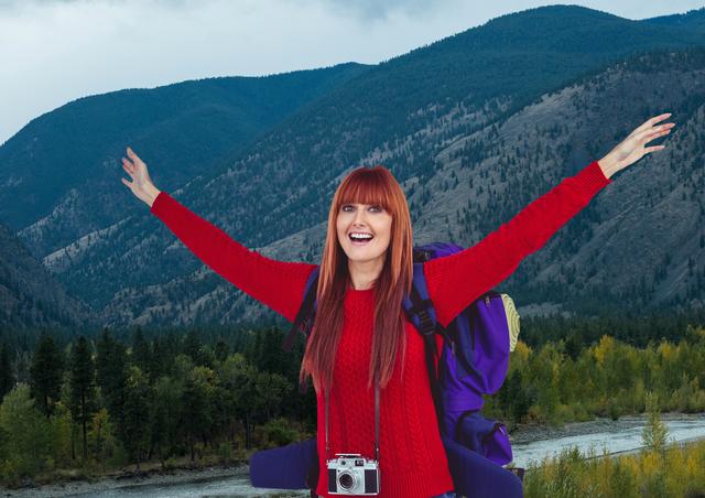 The red-haired woman is smiling broadly, arms outstretched, showcasing enthusiasm and excitement for travel and outdoor adventures. She stands against a backdrop of a mountainous landscape and a flowing river, conveying themes of exploration, happiness, and connection with nature. This image is ideal for camping or travel-themed articles, outdoor adventure blogs, and promotional materials relating to nature hikes and expeditions.