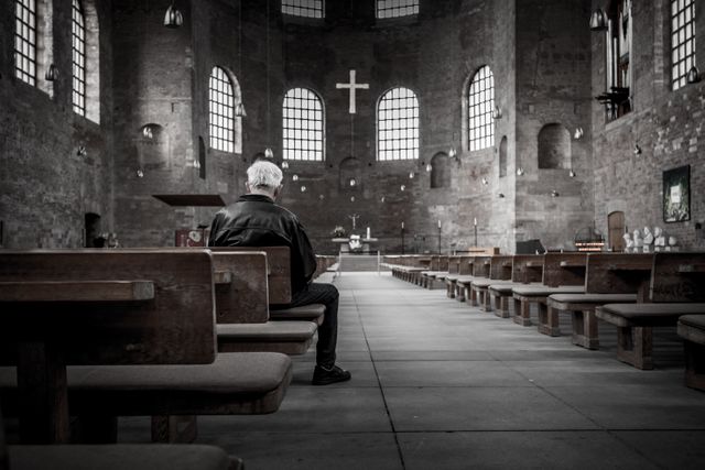 Man sitting alone in large, traditional church interior. Cross in the background emphasizes spirituality and faith. Suitable for concepts like solitude, prayer, worship, architecture, quiet contemplation, and religion.