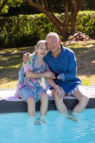Asian senior couple sitting poolside, dipping feet in water, sunlit backyard relaxation