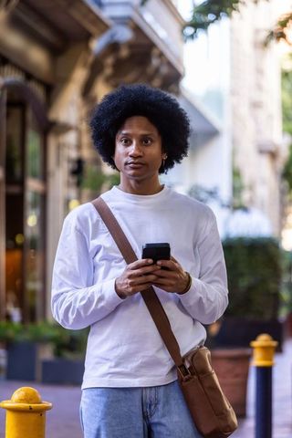 Urban african american man standing on sidewalk holding smartphone wearing crossbody bag