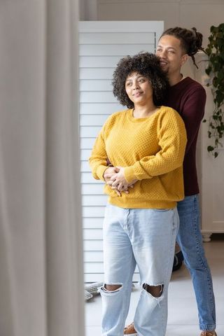 Couple embracing by window in cozy minimalist home wearing knit sweaters and jeans