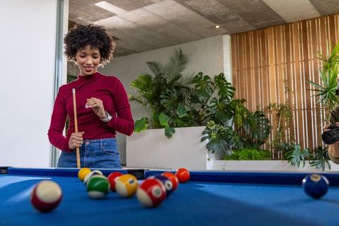 African american woman lining up shot at blue-felt pool table in modern rec room with plants