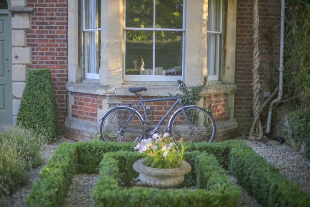 Vintage bicycle leaning against classic brick house featured with an elaborately maintained garden, showcasing neatly trimmed hedges and colorful flowers. Great for use in real estate marketing, gardening, home lifestyle blogs, and vintage equipment articles.