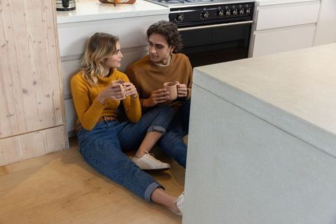 Couple in matching sweaters sits on kitchen floor holding mugs