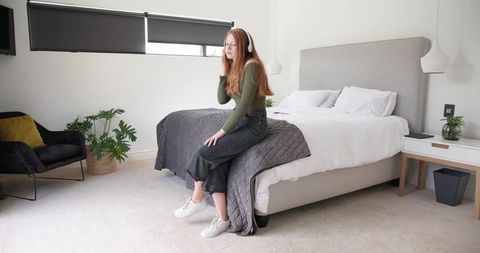 Young woman listening to music on over-ear headphones sitting on bed in minimalist bedroom
