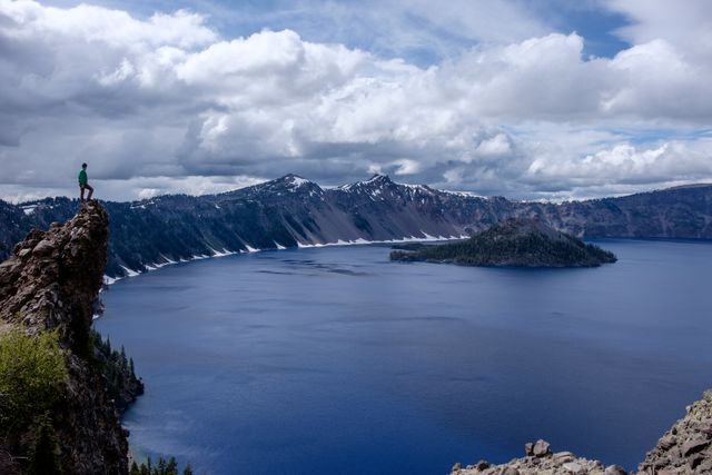 A person stands on a rocky cliff overlooking Crater Lake, with the lake's deep blue water and the surrounding snow-capped mountains under a cloudy sky. Ideal for use in content related to nature, travel, outdoor activities, adventure, scenic viewpoints, or hiking destinations in Oregon.