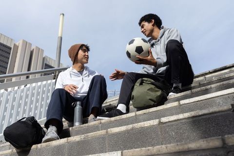 Multicultural teen friends sitting on city steps holding soccer ball and backpack chatting