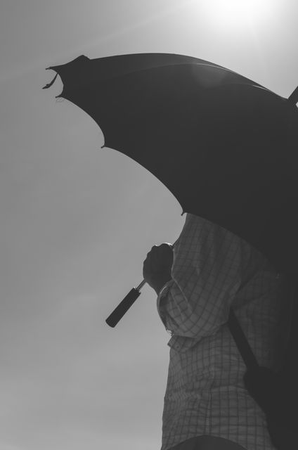 Silhouetted person stands under dark umbrella under bright sun. Ideal for concepts of sun protection, summer heat, walking outdoors, umbrella use, and fashion statement. Perfect for advertisements, promotional materials, storytelling, environmental issues, and editorial content.