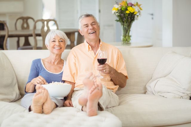 Senior couple sitting on a comfortable couch, enjoying popcorn and a glass of wine. They are smiling and appear relaxed, suggesting a cozy and happy moment at home. This image can be used for promoting senior lifestyle, retirement living, family bonding, and home comfort.