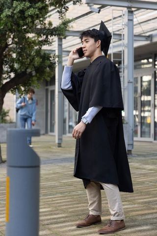 Asian graduate talking on smartphone outside modern university building in cap and gown
