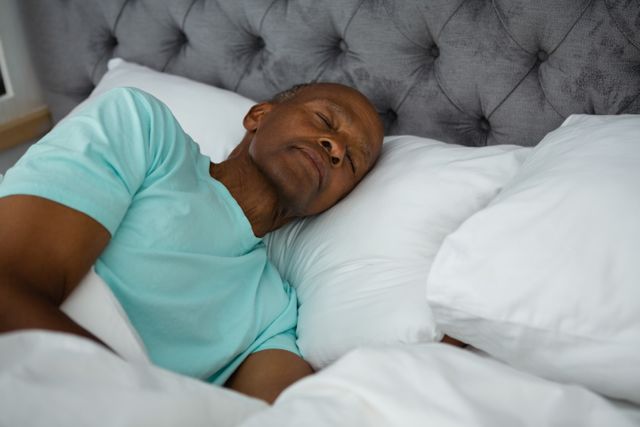 Senior man peacefully sleeping in bed, showcasing comfort and relaxation. Ideal for use in articles or advertisements related to sleep health, elderly care, relaxation techniques, and home comfort products.