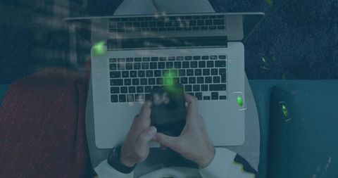 Top-down remote work scene typing on laptop while holding smartphone with green reflection