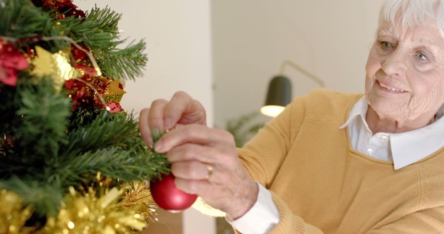 Elderly woman happily hanging a red ornament on a Christmas tree decorated with golden tinsel. Perfect for concepts related to holiday traditions, family gatherings, Christmas celebrations, and festive home decor. Great for use in greeting cards, holiday advertisements, and articles on family celebrations.