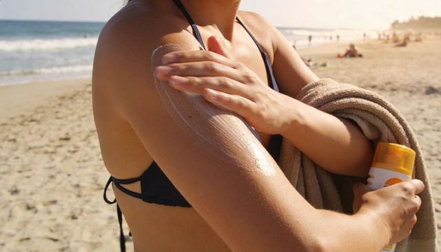Woman applying spf50 sunscreen on arm at sunny beach holding towel in bikini