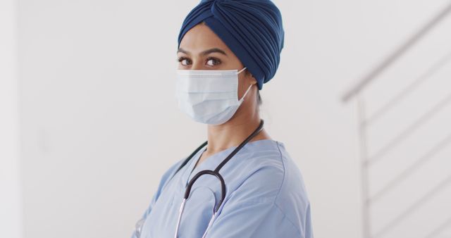 Confident healthcare worker wearing turban and protective mask while standing with crossed arms, stethoscope around neck. Ideal for use in medical articles, hospital promotional materials, healthcare hero campaigns, COVID-19 safety and awareness graphics.