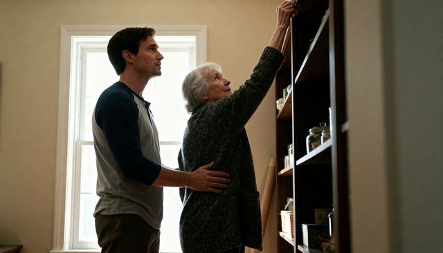 Adult son supporting elderly mother reaching high pantry shelf for home caregiving support