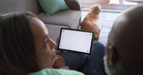 Diverse senior couple relaxing on sofa holding tablet with blank screen and dog nearby