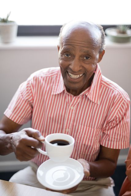 Senior man sitting indoors, smiling while holding a cup of coffee. Ideal for use in materials related to elderly care, retirement living, happiness in old age, and lifestyle content for seniors.