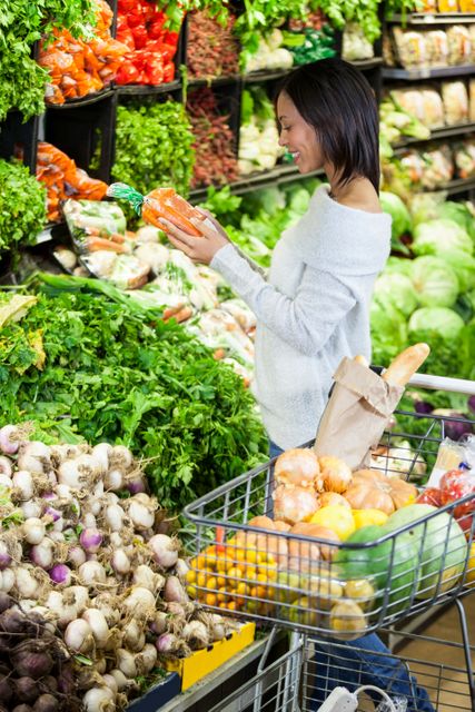 Woman selecting fresh organic carrots in supermarket produce section. Shopping cart filled with various groceries, including fruits and bread. Ideal for illustrating healthy lifestyle, grocery shopping, organic food choices, and retail consumer behavior.