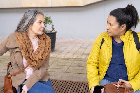 Two mature women having thoughtful conversation on bench in urban plaza wearing jackets