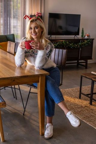 Young woman wearing festive sweater holding red mug and smiling with candy-cane headband