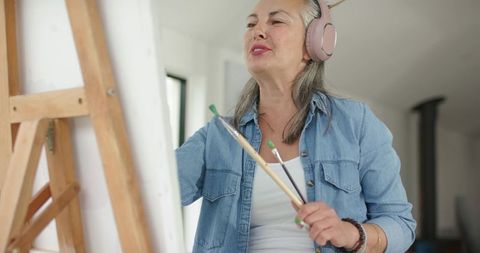 Mature woman painting on canvas wearing headphones and denim shirt in bright studio