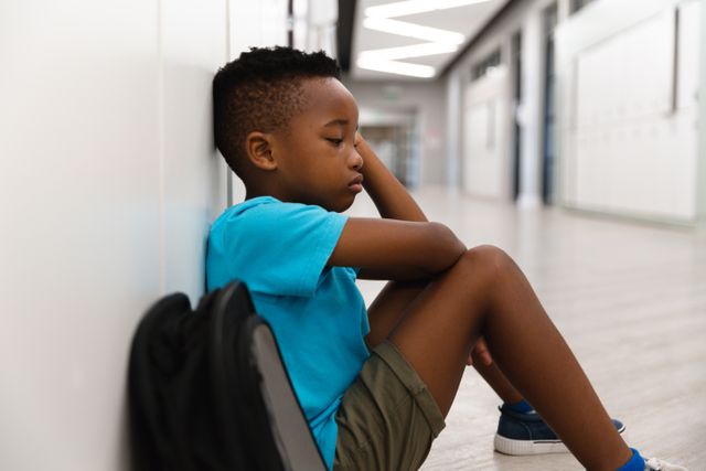 Side view of sad african american elementary schoolboy sitting lonely on floor in corridor. unaltered, childhood, education, sadness, loneliness, failure and school concept.