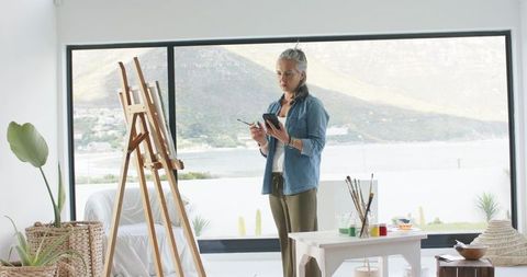 Senior woman painting by waterfront studio standing at easel with smartphone and brushes