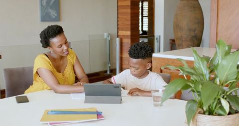 African American mother helping son with homework using tablet at modern dining table, home learning