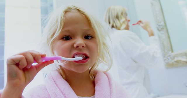 Child brushing her teeth with pink toothbrush in bathroom while parent is brushing in background. Example of teaching cleanliness and healthy habits to children. Ideal for promoting dental hygiene, family routines and parental education.
