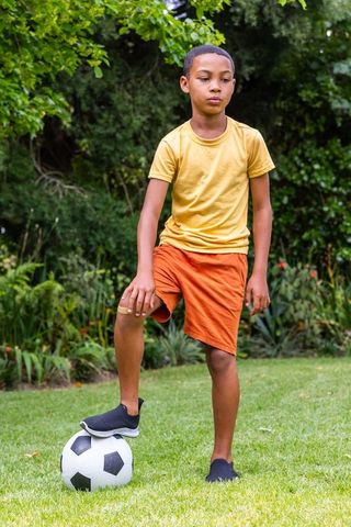 African american boy balancing on soccer ball in backyard garden wearing knee bandage