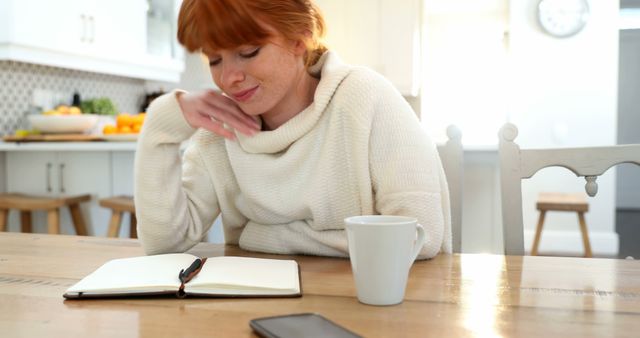 Redhead woman sits at a wooden table in her home kitchen, journaling quietly with a white coffee mug nearby. Light floods the room through large windows, adding to a cozy and serene atmosphere, reinforced by a white sweater. This image is ideal for use in lifestyle blogs, morning routine promotions, or mental health and self-care discussions.