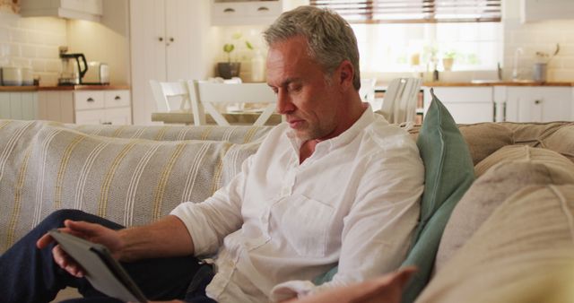 Middle-aged man wearing a casual white shirt, sitting comfortably on a couch while using a digital tablet in a bright and modern living room with natural light. White and beige decor create a cozy atmosphere. Ideal for use in articles about technology use, home leisure activities, modern living, aging population and technology, and lifestyle content.