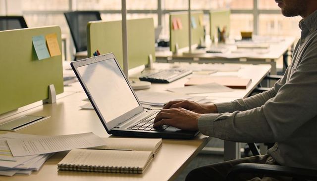 Typing professional working at open-plan office desk with laptop, notebooks, paperwork